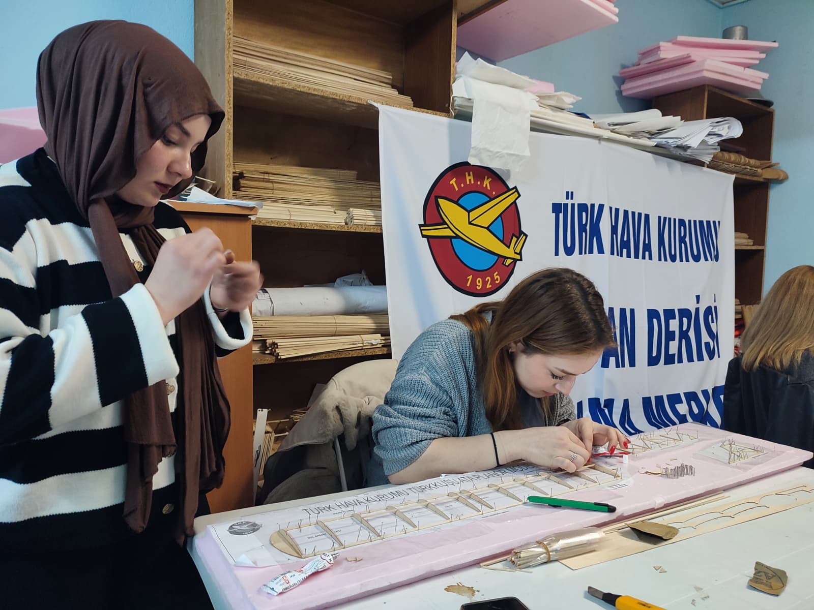 Öğrencilerin atölyede model uçak yapımıyla ilgilendiklerini gösteriyor. Öğrenciler, eğitmenlerin yardımıyla uçak parçalarını bir araya getiriyor, yapıştırıyor ve şekillendiriyor. Atölye ortamı, öğrencilerin öğrenmeye istekli ve keyifli bir şekilde çalıştıklarını yansıtıyor.